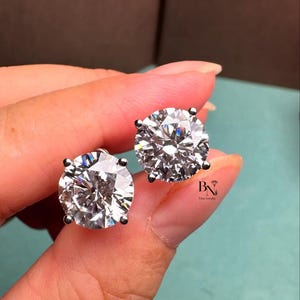 Peut inclure: Une paire de boucles d'oreilles à clous en diamant rond taille brillant. Les diamants sont sertis dans un métal de couleur argentée. Les boucles d'oreilles sont présentées sur un fond bleu clair. Bijoux fins BN.