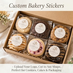 May include: A cardboard box filled with six individually wrapped cookies. Each cookie has a round sticker that says "Wildflower Bakery". The cookies are various flavors, including chocolate chip and frosted sugar cookies. The text "Custom Bakery Stickers" is at the top.