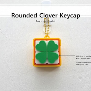May include: A square, yellow and white keycap with four green clover leaves. The keycap is suspended by a gold chain. The text "Rounded Clover Keycap" is at the top of the image.