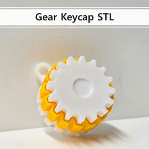 Könnte beinhalten: Ein 3D-gedruckter Gear-Keycap in Gelb und Weiß. Der Keycap hat ein Zahnraddesign mit einem weißen Zentrum und einer gelben Außenschicht. Der Text "Gear Keycap STL" befindet sich darüber.