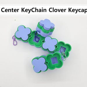 Peut inclure: Un porte-clés en forme de trèfle, de couleur violet clair et vert. Le porte-clés est composé de plusieurs pièces en forme de trèfle reliées entre elles, avec une petite chaîne et une perle violette. Le texte "Left Center KeyChain Clover Keycap STL" est en haut.