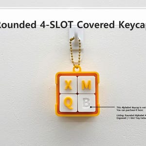 May include: A yellow and white keycap with the letters X, M, Q, and B. The keycap is square with rounded corners and is attached to a gold chain. The text "Rounded 4-SLOT Covered Keycap" is above the keycap.
