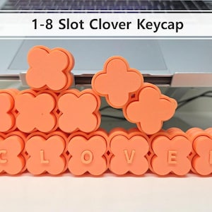 Puede incluir: Teclas naranjas en forma de trébol que deletrean la palabra "CLOVER". Las teclas están apiladas frente a una computadora portátil. El texto "1-8 Slot Clover Keycap" se muestra encima de las teclas.