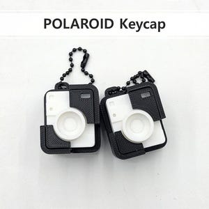 May include: Two Polaroid keycaps, each featuring a black and white design resembling a classic instant camera. The keycaps have a circular lens and are attached to a small chain.