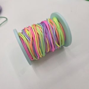May include: A spool of colorful cord in shades of pink, yellow, orange, purple, and green. The cord is tightly wound around a light blue spool, creating a vibrant and eye-catching display. Ideal for crafting and jewelry making.