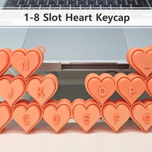May include: A collection of coral-colored heart-shaped keycaps, each with a letter of the alphabet. The keycaps are arranged in rows and columns, with the text "1-8 Slot Heart Keycap" above them. The keycaps are likely for a keyboard.