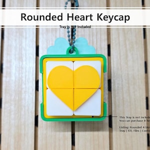 May include: A green and yellow keycap with a rounded heart design. The keycap features a yellow heart shape in the centre, surrounded by white and green squares. The text "Rounded Heart Keycap" is above the keycap.
