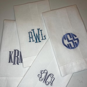 Può includere: Una collezione di tovaglioli di lino bianco, ciascuno ricamato con un monogramma diverso in tonalità di blu. I monogrammi variano nello stile, tra cui lettere a blocchi e un design circolare. I tovaglioli sono piegati e disposti con cura.