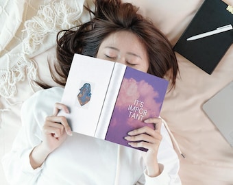 Op de afbeelding: Een boek met een paarse kaft met een wolkenafbeelding en de tekst IT'S IMPORTANT TANT, opengehouden om een witte pagina met een kleine blauwe en oranje sticker te tonen. Het boek ligt over een gezicht, vergezeld door een zwart notitieboek en een pen.