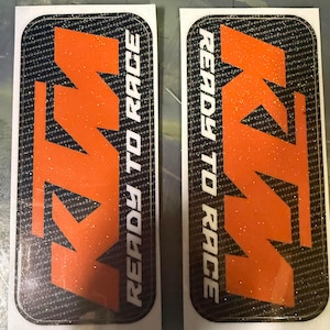 May include: Two rectangular stickers with a carbon fiber pattern background. The stickers feature the KTM logo in orange, with the words "READY TO RACE" in white. The stickers have a glittery finish.