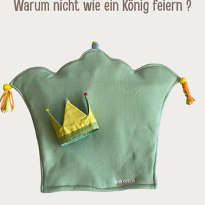 May include: A sage green crown-shaped pillow with yellow and orange tassels on the sides, featuring a small yellow fabric crown placed on top. The text "Warum nicht wie ein K&ouml;nig feiern ?" is at the top and "anki kids" is at the bottom.