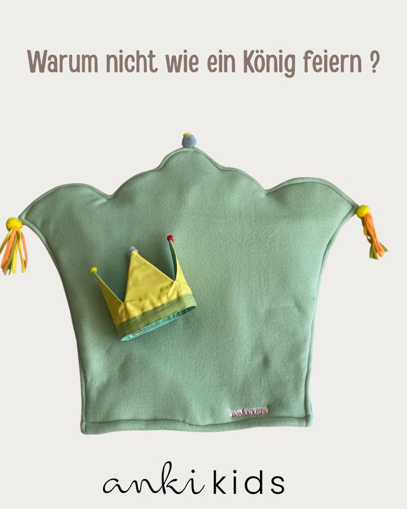 May include: A sage green crown-shaped pillow with yellow and orange tassels on the sides, featuring a small yellow fabric crown placed on top. The text "Warum nicht wie ein K&ouml;nig feiern ?" is at the top and "anki kids" is at the bottom.