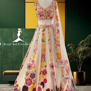 May include: A cream-colored lehenga choli set featuring a sleeveless blouse and a floor-length skirt adorned with vibrant, multi-colored floral embroidery in shades of pink, yellow, and purple. A matching sheer dupatta with mirror-work borders.