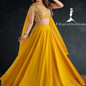 Puede incluir: Un conjunto de dos piezas amarillo vibrante. La parte superior es un diseño corto y sin mangas con intrincados bordados en tonos amarillos, verdes y naranjas. La falda es un estilo largo y fluido. Una bufanda y un collar a juego completan el conjunto.