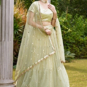 May include: A light green lehenga set featuring a short-sleeved blouse and a floor-length skirt, both adorned with intricate gold embroidery and sequins. A matching sheer dupatta with a gold-trimmed border drapes over the shoulders.
