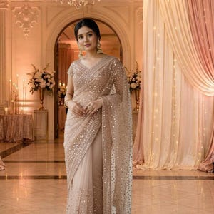 Może przedstawiać: Beżowe sari z misternymi detalami z cekinów. Przezroczysty materiał układa się elegancko, tworząc efekt połysku. Sari jest noszone z pasującą bluzką. Tło przedstawia żyrandol i elegancki wystrój.