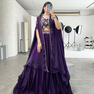 Puede incluir: Conjunto formal morado oscuro con falda de capas, top corto bordado y capa fluida. El atuendo se complementa con un delicado collar de perlas, adecuado para ocasiones especiales.