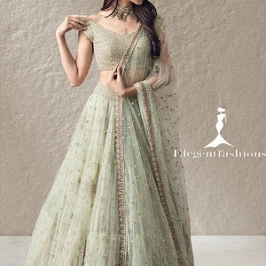 Op de afbeelding: Een lichtgroene lehenga choli-set met ingewikkeld borduurwerk en paillettenversieringen. Het ensemble bestaat uit een blouse met korte mouwen, een volumineuze rok tot op de grond en een transparante dupatta. De tekst Elegant fashions is zichtbaar.