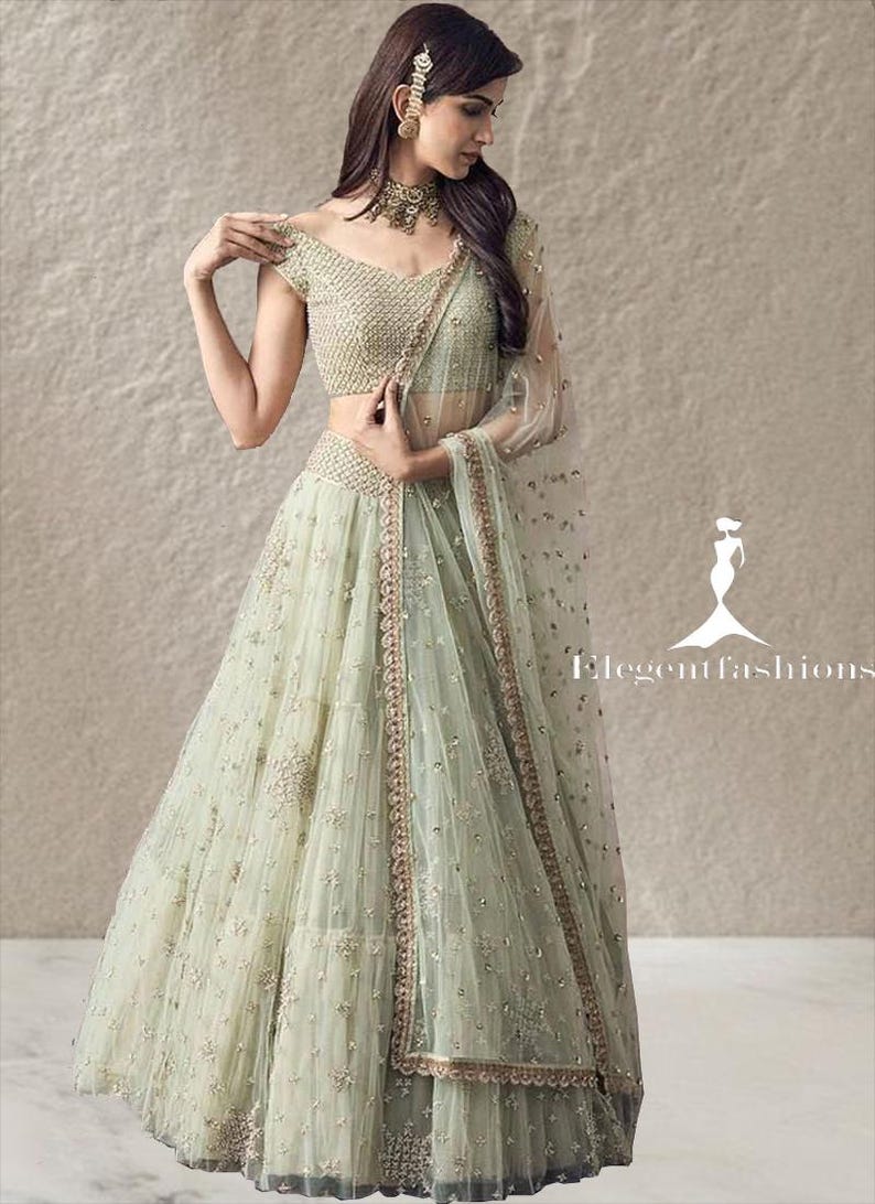 Op de afbeelding: Een lichtgroene lehenga choli-set met ingewikkeld borduurwerk en paillettenversieringen. Het ensemble bestaat uit een blouse met korte mouwen, een volumineuze rok tot op de grond en een transparante dupatta. De tekst Elegant fashions is zichtbaar.