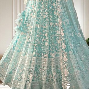 May include: A light blue formal gown with intricate white floral embroidery. The gown features a full skirt with detailed patterns and a matching sheer overlay. The design includes a ruffled accent and a flowing, sheer scarf.