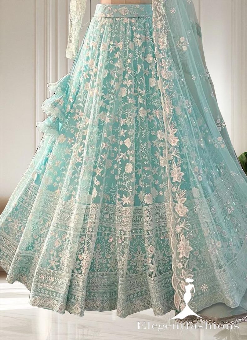 May include: A light blue formal gown with intricate white floral embroidery. The gown features a full skirt with detailed patterns and a matching sheer overlay. The design includes a ruffled accent and a flowing, sheer scarf.