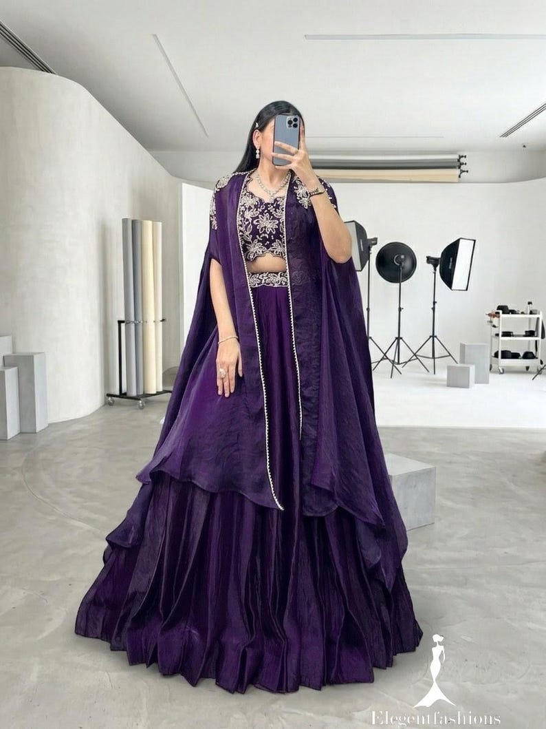 Puede incluir: Conjunto formal morado oscuro con falda de capas, top corto bordado y capa fluida. El atuendo se complementa con un delicado collar de perlas, adecuado para ocasiones especiales.