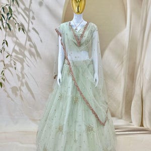 Op de afbeelding: Een licht mintgroene lehenga choli-set bestaande uit een mouwloze blouse en een vloerlange rok van doorschijnende stof. Het kledingstuk is versierd met verspreide gouden lovertjes en borduursels, met een bijpassende dupatta met een rand.