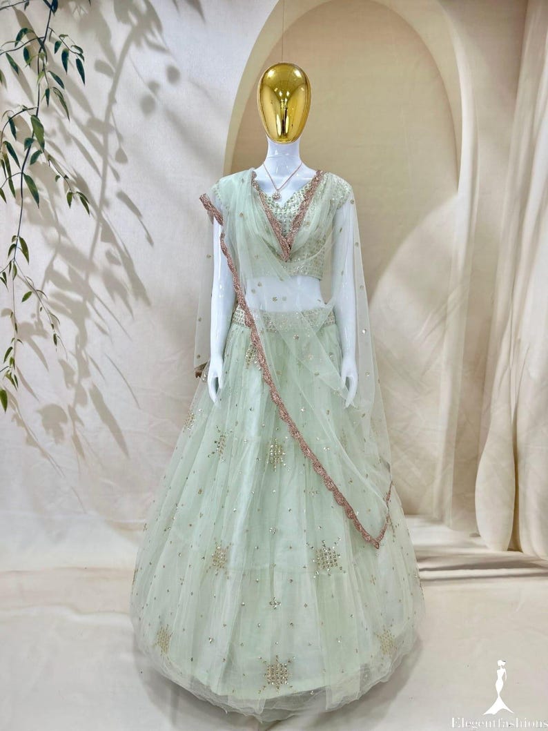 Op de afbeelding: Een licht mintgroene lehenga choli-set bestaande uit een mouwloze blouse en een vloerlange rok van doorschijnende stof. Het kledingstuk is versierd met verspreide gouden lovertjes en borduursels, met een bijpassende dupatta met een rand.