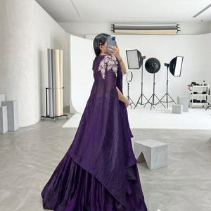 Puede incluir: Un vestido largo y fluido de color p&uacute;rpura oscuro con una falda en capas y una capa a juego. La capa est&aacute; adornada con bordados florales blancos en los hombros. El vestido est&aacute; hecho de una tela brillante y el estilo general es elegante.