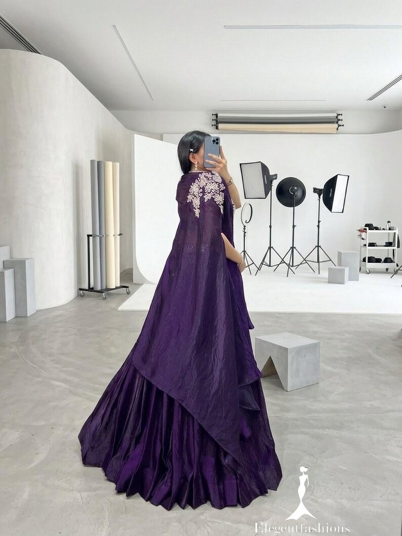 Puede incluir: Un vestido largo y fluido de color p&uacute;rpura oscuro con una falda en capas y una capa a juego. La capa est&aacute; adornada con bordados florales blancos en los hombros. El vestido est&aacute; hecho de una tela brillante y el estilo general es elegante.
