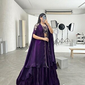 Puede incluir: Un conjunto formal de color p&uacute;rpura oscuro, con una falda en capas y una parte superior estilo capa con bordados intrincados. El atuendo incluye un top corto a juego y est&aacute; dise&ntilde;ado para ocasiones especiales. La imagen se toma en un estudio.