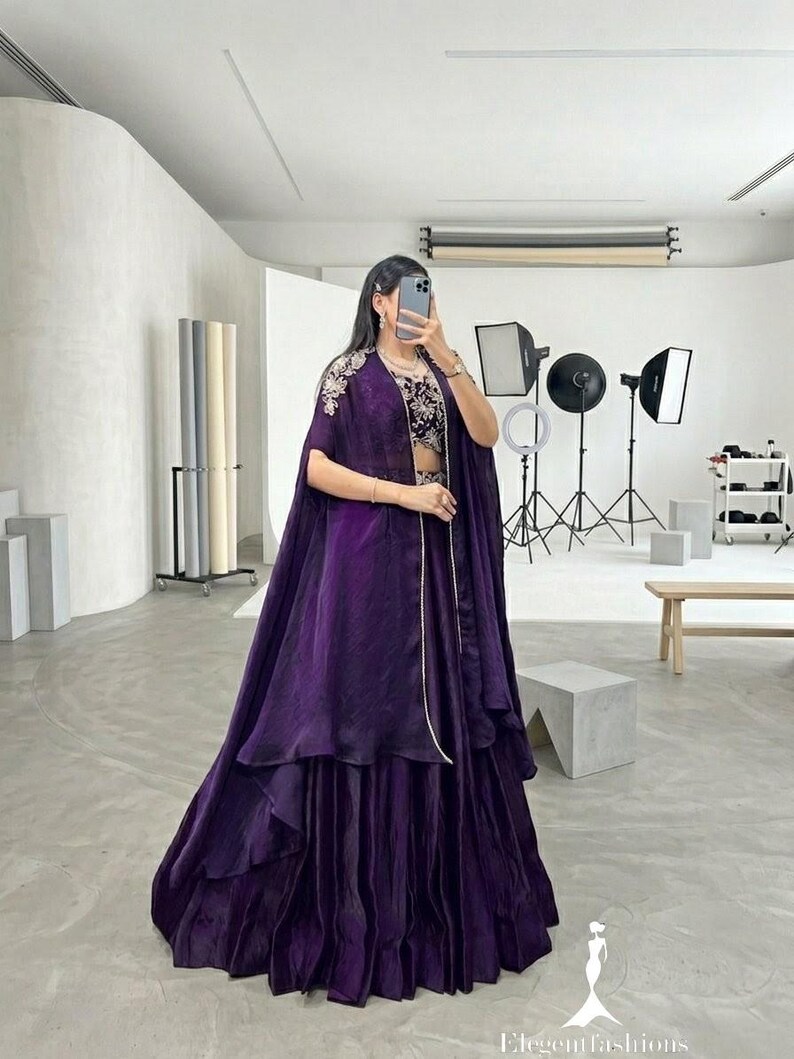 Puede incluir: Un conjunto formal de color p&uacute;rpura oscuro, con una falda en capas y una parte superior estilo capa con bordados intrincados. El atuendo incluye un top corto a juego y est&aacute; dise&ntilde;ado para ocasiones especiales. La imagen se toma en un estudio.