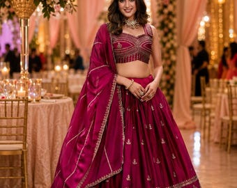 May include: A woman wearing a magenta-colored, embroidered lehenga choli, a traditional Indian outfit. The outfit includes a fitted, embellished top and a flowing skirt with gold detailing. She is wearing a matching dupatta and jewelry. The background features a decorated event space.