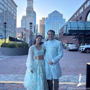 May include: A couple poses outdoors, dressed in matching light blue formal wear. The woman wears a crop top and skirt with lace sleeves. The man wears a long jacket with ornate detailing. The background features city buildings and a yellow bus.