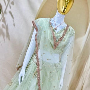 Op de afbeelding: Een licht mintgroene lehenga choli-set met een transparante dupatta met geschulpte ros&eacute;gouden borduursels. De blouse en rok zijn versierd met ingewikkeld paillettenwerk en metalen accenten. Een goudkleurige ketting wordt gedragen door de etalagepop.