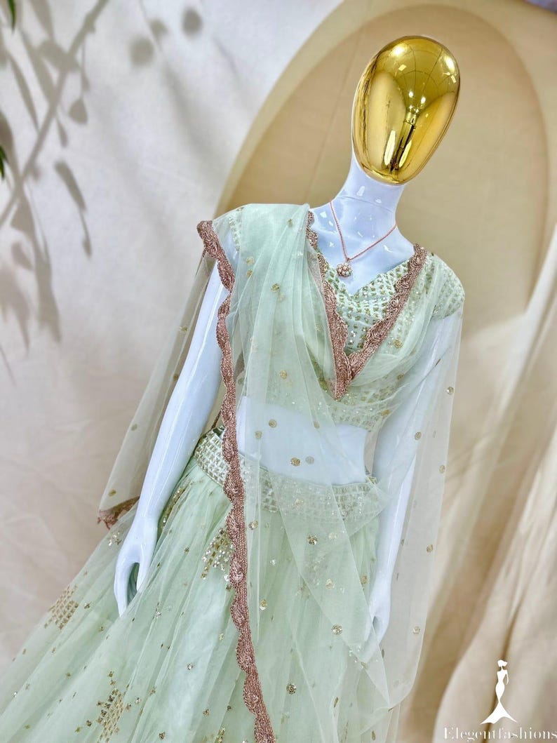Op de afbeelding: Een licht mintgroene lehenga choli-set met een transparante dupatta met geschulpte ros&eacute;gouden borduursels. De blouse en rok zijn versierd met ingewikkeld paillettenwerk en metalen accenten. Een goudkleurige ketting wordt gedragen door de etalagepop.