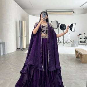 Puede incluir: Un conjunto formal de color p&uacute;rpura oscuro que presenta un top corto bordado, una falda a juego y una capa fluida. El conjunto se complementa con un delicado collar de perlas. La imagen est&aacute; tomada en un estudio.