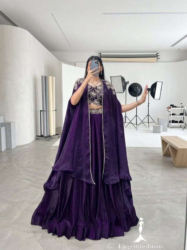 Puede incluir: Un conjunto formal de color p&uacute;rpura oscuro que presenta un top corto bordado, una falda a juego y una capa fluida. El conjunto se complementa con un delicado collar de perlas. La imagen est&aacute; tomada en un estudio.