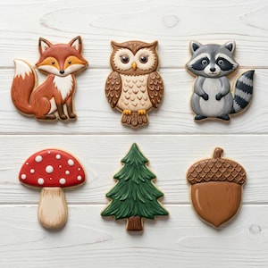 May include: A collection of six woodland-themed cookies. The cookies include a fox, owl, raccoon, mushroom, pine tree, and acorn. The cookies are decorated with brown, red, white, and green icing and are arranged on a white wooden surface.