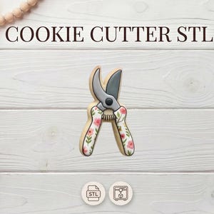 May include: A cookie cutter shaped like garden shears with a floral design on the handles. The shears are gray and black. The text "COOKIE CUTTER STL" is at the top of the image. The background is a white wooden surface.