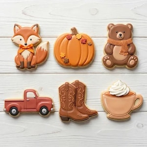 May include: A collection of decorated sugar cookies in autumn themes. The cookies include a fox, a pumpkin, a bear with a scarf, a pickup truck, cowboy boots, and a coffee cup with whipped cream. The cookies are on a white wooden surface.