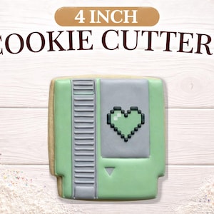 May include: A 4-inch cookie cutter shaped like a retro video game cartridge. The cookie is decorated with light green, gray, and black icing, featuring a pixelated heart design. The words "COOKIE CUTTERS" are above the cookie.