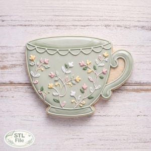 Può includere: Un biscotto a forma di tazza da tè verde chiaro decorato con motivi floreali in glassa bianca, gialla e rosa. Il biscotto ha un bordo smerlato e un manico. Il testo "STL File" si trova nell'angolo in basso a sinistra.