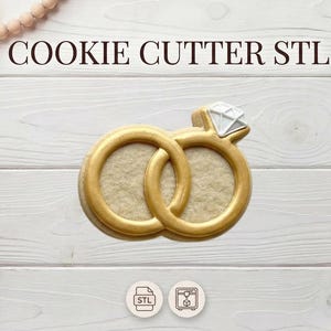 Puede incluir: Cortador de galletas dorado en forma de dos anillos entrelazados, uno con una gema en forma de diamante. El texto "COOKIE CUTTER STL" está en la parte superior. El cortador está sobre una superficie de madera blanca.