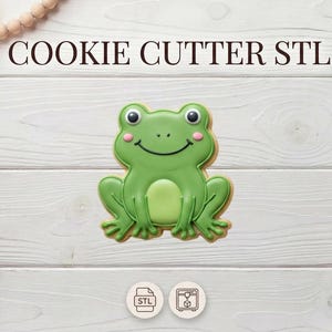 Peut inclure: Un emporte-pièce en forme de grenouille verte avec un visage souriant et des joues roses. Les mots "COOKIE CUTTER STL" sont en haut. L'emporte-pièce est sur une surface en bois blanc avec des éléments décoratifs.
