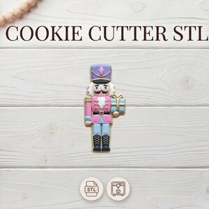 May include: A colorful cookie cutter in the shape of a nutcracker soldier, holding a gift. The nutcracker is pink, blue, and purple, with black boots. The text "COOKIE CUTTER STL" is above the cookie cutter.
