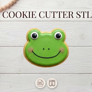 Könnte beinhalten: Ein grüner, froschförmiger Keks mit einem lächelnden Gesicht, schwarzen Augen und beigen Akzenten. Der Keks liegt auf einer weißen Holzoberfläche. Der Text "COOKIE CUTTER STL" steht oben.