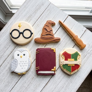 May include: A set of six decorated sugar cookies featuring a round face with black glasses and a yellow lightning bolt, a brown pointed hat, a light brown wand, a white owl, a rectangular maroon book with gold corners, and a red and green shield.