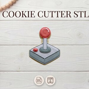 Puede incluir: Un cortador de galletas con forma de joystick de videojuego retro. El joystick tiene una bola roja en la parte superior y un botón rojo en la base. El texto "COOKIE CUTTER STL" está en la parte superior.