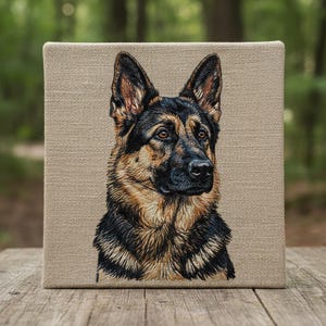 Puede incluir: Retrato bordado de un perro pastor alemán sobre un lienzo cuadrado. El pelaje del perro está representado en hilos negros, marrones y beige, con una apariencia detallada y realista. El lienzo se muestra sobre un fondo natural borroso.